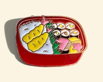 Op de afbeelding: Een rode emaille speld in de vorm van een bentobox met witte rijst, twee gele gefrituurde garnalen, groene garnering, diverse donkerbruine sushirolletjes, een roze stukje vis en een geel vierkant met een roze ster, afgewerkt met koperen randen.