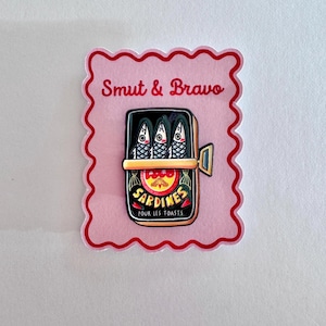 May include: A rectangular enamel pin featuring an open tin of three sardines with stylized faces. The pin is mounted on a light pink backing card with a scalloped red border and the text Smut & Bravo printed in red at the top.