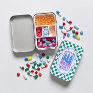 Peut inclure: Un pilulier rectangulaire en métal avec un couvercle à charnière, orné d'un motif à damier vert et blanc et d'un dessin de sardines en boîte avec le texte SARDINES. Le récipient ouvert contient diverses gélules et comprimés colorés.