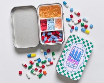 Puede incluir: Un pastillero rectangular de metal con tapa abatible que presenta un patrón de tablero de ajedrez verde y blanco y un gráfico de tres sardinas con la etiqueta SARDINES. El recipiente abierto contiene varias cápsulas y pastillas.