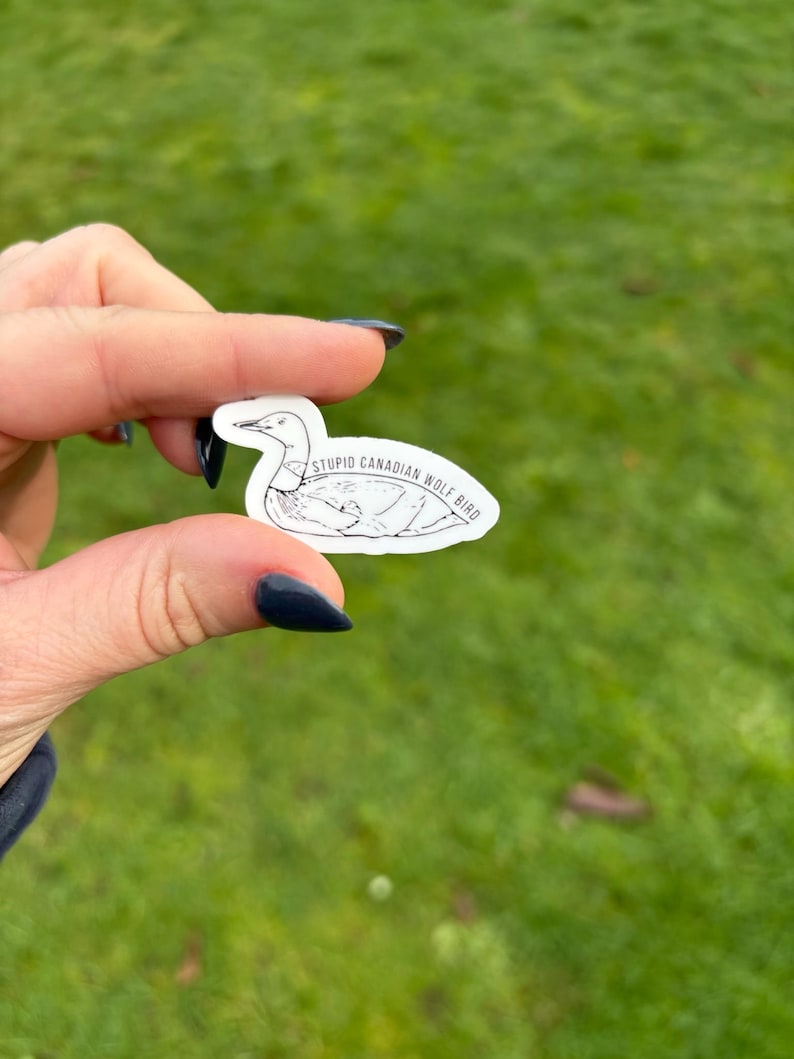 K&ouml;nnte beinhalten: Ein wei&szlig;er Aufkleber mit einer schwarzen Umrisszeichnung eines Vogels und dem Text "STUPID CANADIAN WOLF BIRD". Der Aufkleber wird zwischen zwei Fingern vor einem gr&uuml;nen Gras Hintergrund gehalten.