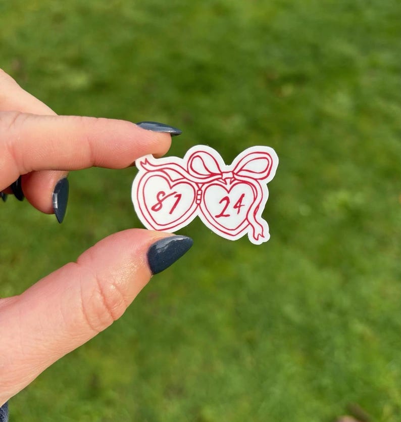 K&ouml;nnte beinhalten: Ein wei&szlig;er Aufkleber mit roter Umrandung, der zwei verbundene herzf&ouml;rmige Designs zeigt. Jedes Herz enth&auml;lt die Zahlen "81" und "24". Eine rote Schleife verbindet die Herzen. Der Aufkleber wird vor einem gr&uuml;nen Hintergrund gehalten.