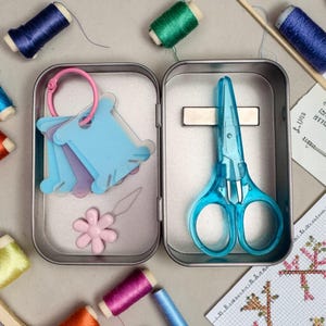 May include: A metal tin containing a pair of translucent blue scissors, a pink flower-shaped needle threader, and a set of light blue plastic thread bobbins on a pink ring. Several spools of colorful embroidery thread surround the open tin.