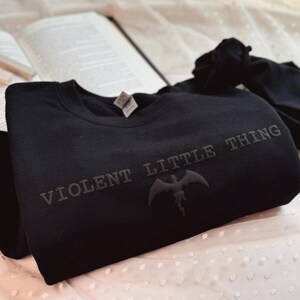 Puede incluir: Sudadera negra con el texto "VIOLENT LITTLE THING" y un gr&aacute;fico de murci&eacute;lago. La sudadera est&aacute; hecha de un material suave y se muestra sobre una superficie blanca con lunares, con un libro en el fondo.