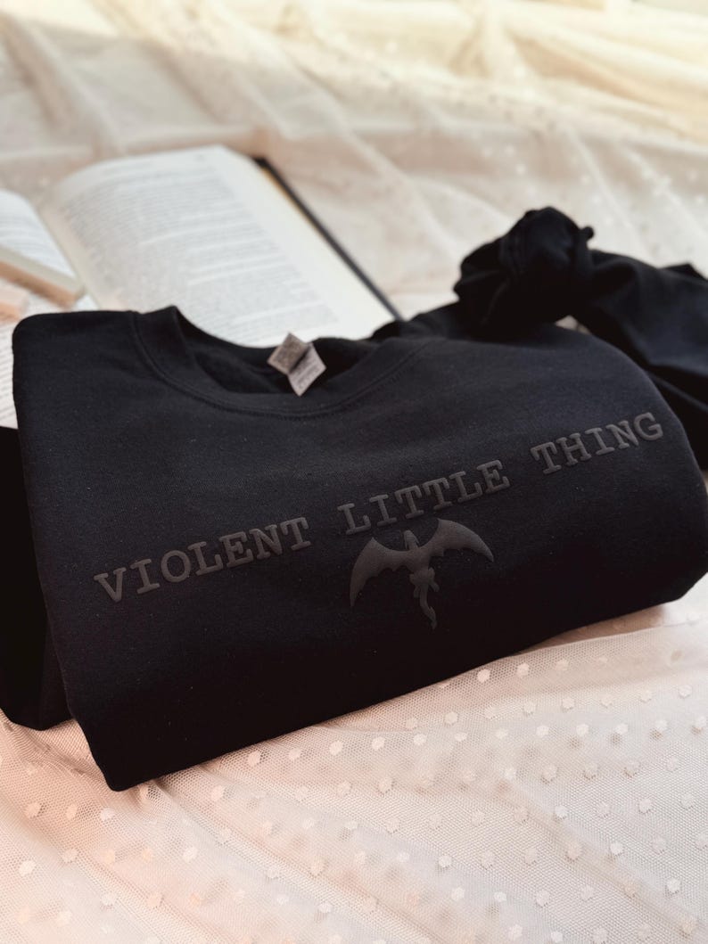 Puede incluir: Sudadera negra con el texto "VIOLENT LITTLE THING" y un gr&aacute;fico de murci&eacute;lago. La sudadera est&aacute; hecha de un material suave y se muestra sobre una superficie blanca con lunares, con un libro en el fondo.