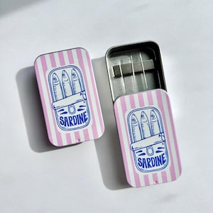 May include: Two rectangular pink and white striped tins, one open, with a blue illustration of sardines in a can. The word "SARDINE" is printed in blue below the illustration. The tins are on a white surface.