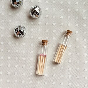 May include: Two small glass bottles filled with matches on a white background with silver disco balls.