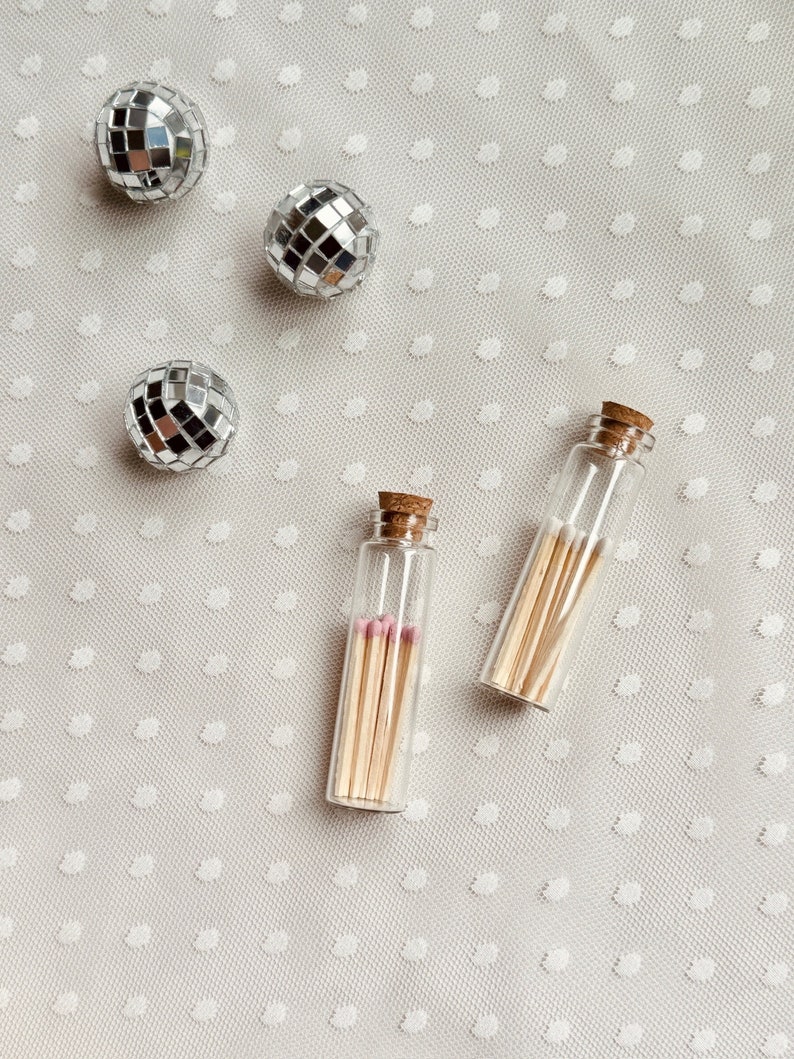 May include: Two small glass bottles filled with matches on a white background with silver disco balls.