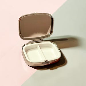 May include: A small, rectangular, silver-toned metal pill case with a hinged lid. The interior features a reflective mirror on the inside of the lid and a white plastic insert divided into two separate compartments for storing small items.