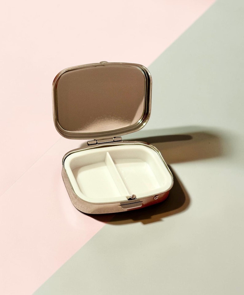 May include: A small, rectangular, silver-toned metal pill case with a hinged lid. The interior features a reflective mirror on the inside of the lid and a white plastic insert divided into two separate compartments for storing small items.