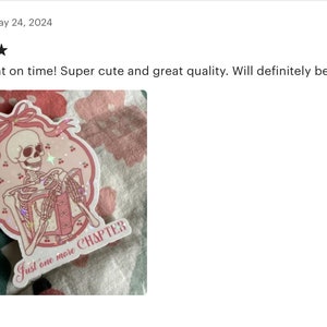 May include: Pink and white sticker with a skeleton holding a book. The sticker has a pink ribbon and the text "Just one more CHAPTER".