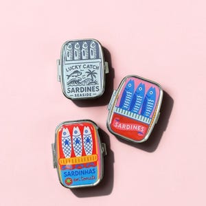 May include: Three rectangular metal tins with hinged lids, each featuring a colorful graphic of sardines. One tin is blue with text reading Lucky Catch Sardines Seaside, one is pink with blue fish, and one is red with text Sardinhas em tomate.