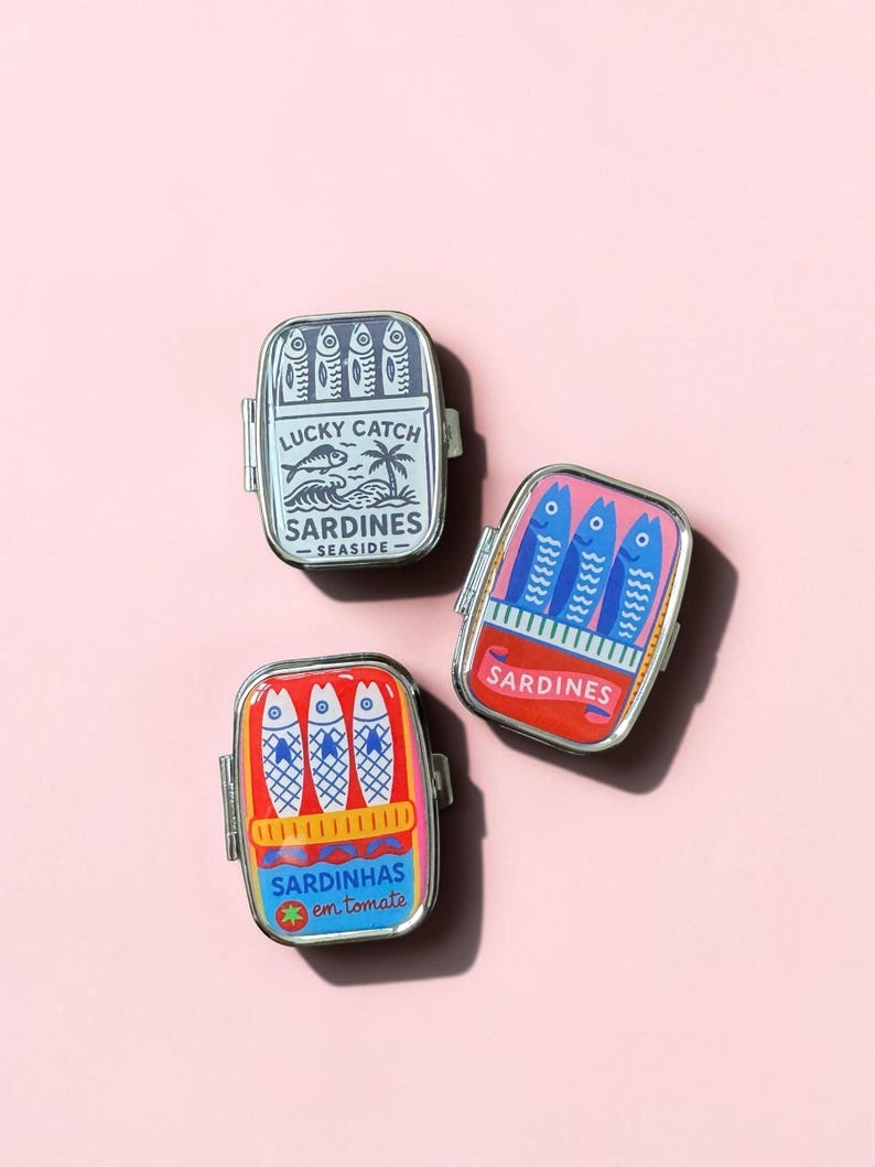 May include: Three rectangular metal tins with hinged lids, each featuring a colorful graphic of sardines. One tin is blue with text reading Lucky Catch Sardines Seaside, one is pink with blue fish, and one is red with text Sardinhas em tomate.