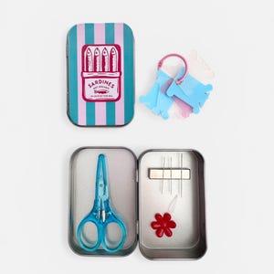 May include: A rectangular metal tin with a teal and pink striped lid featuring a graphic of four sardines and the text SARDINES NET DREAMS IN LOVE BY THE SEA. Inside are blue scissors, three needles, a red flower-shaped needle threader, and thread bobbins.