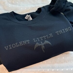 Puede incluir: Una sudadera negra con el texto "VIOLENT LITTLE THING" y un gr&aacute;fico de drag&oacute;n. La sudadera est&aacute; hecha de un material suave y est&aacute; doblada sobre una superficie blanca con lunares.