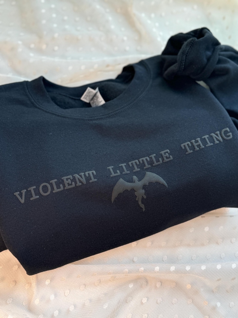 Puede incluir: Una sudadera negra con el texto "VIOLENT LITTLE THING" y un gr&aacute;fico de drag&oacute;n. La sudadera est&aacute; hecha de un material suave y est&aacute; doblada sobre una superficie blanca con lunares.