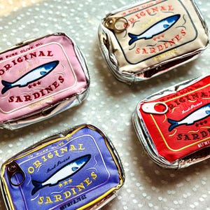 May include: Four sardine-shaped pouches in pink, tan, red, and blue. Each pouch features a sardine illustration and the words "Original Sardines" in a vintage style. The pouches are labeled with the net weight in ounces.