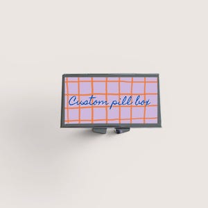 Op de afbeelding: Een rechthoekige pillendoos met een lavendelkleurig rasterpatroon en de woorden "Custom pill box" in blauw schrift. De rasterlijnen zijn oranje. De doos heeft een zilverkleurig metalen frame en een kleine sluiting.