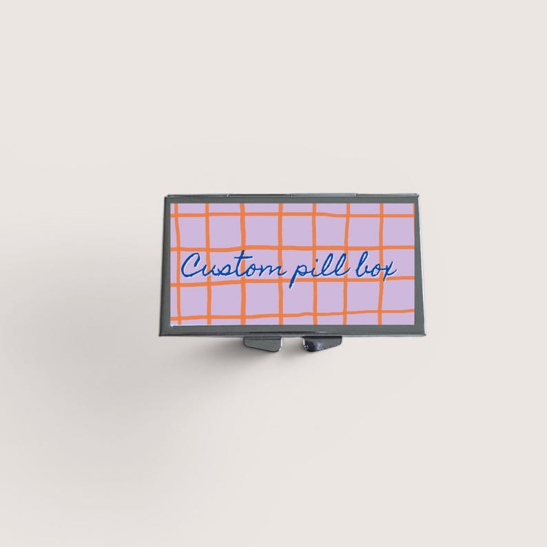 Op de afbeelding: Een rechthoekige pillendoos met een lavendelkleurig rasterpatroon en de woorden "Custom pill box" in blauw schrift. De rasterlijnen zijn oranje. De doos heeft een zilverkleurig metalen frame en een kleine sluiting.