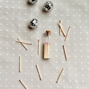 May include: A glass bottle filled with wooden matches with pink tips, surrounded by scattered matches on a white background with a polka dot pattern. Three silver disco balls are also scattered on the background.