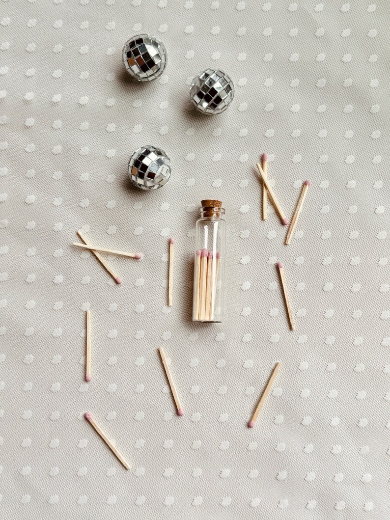 May include: A glass bottle filled with wooden matches with pink tips, surrounded by scattered matches on a white background with a polka dot pattern. Three silver disco balls are also scattered on the background.
