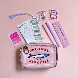 May include: A pink rectangular pouch featuring an embroidered sardine tin design with the text Original Sardines and Net Wt 3.75 oz. Includes a reading list card, a stack of books bookmark with a tassel, two light purple pens, and three stickers.