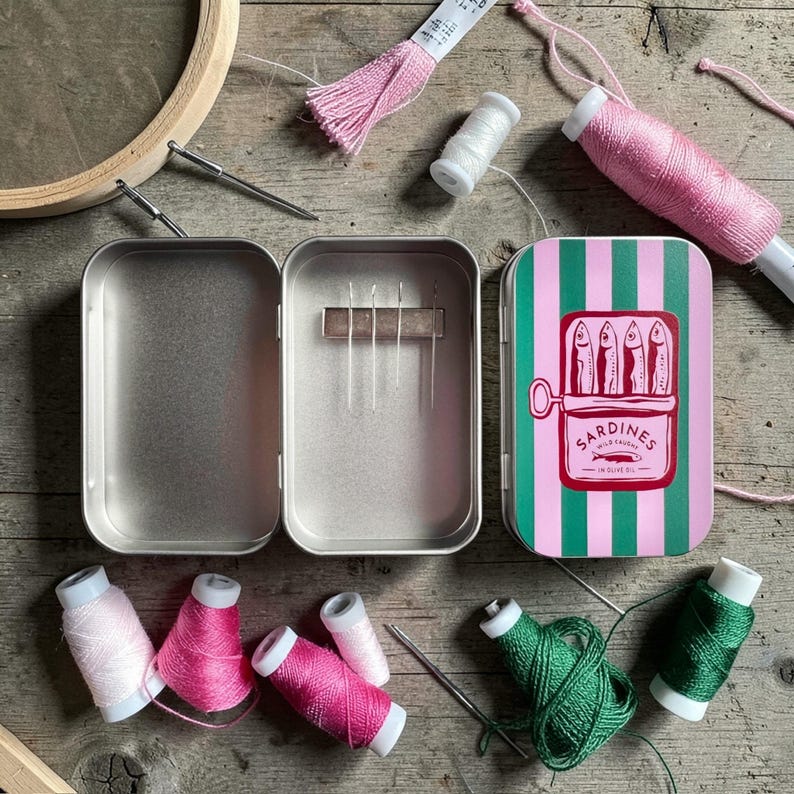 May include: A metal sewing needle case with a hinged lid featuring a pink and green striped sardine tin graphic. The interior contains a magnetic strip holding four needles. Surrounding the case are various spools of pink, white, and green thread.