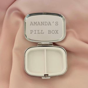 K&ouml;nnte beinhalten: Ge&ouml;ffnete, silberfarbene Pillendose mit dem Text "AMANDA'S PILL BOX" auf dem Deckel. Der Innenraum ist in zwei F&auml;cher unterteilt. Die Pillendose befindet sich vor einem weichen, rosa Hintergrund.