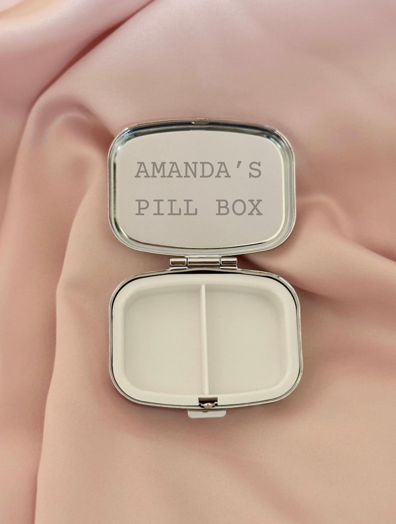 K&ouml;nnte beinhalten: Ge&ouml;ffnete, silberfarbene Pillendose mit dem Text "AMANDA'S PILL BOX" auf dem Deckel. Der Innenraum ist in zwei F&auml;cher unterteilt. Die Pillendose befindet sich vor einem weichen, rosa Hintergrund.