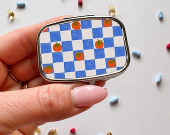 Puede incluir: Un pastillero rectangular plateado con un patrón de cuadros azules y blancos e ilustraciones de tomates naranjas. El estuche se sostiene en una mano, rodeado de varias pastillas de diferentes colores.