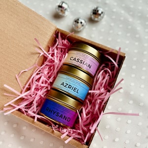 May include: Three gold candle tins with pink, blue, and purple labels. The labels have the names "Cassian", "Azriel", and "Rhysand" on them. The candles are in a brown box with pink shredded paper.