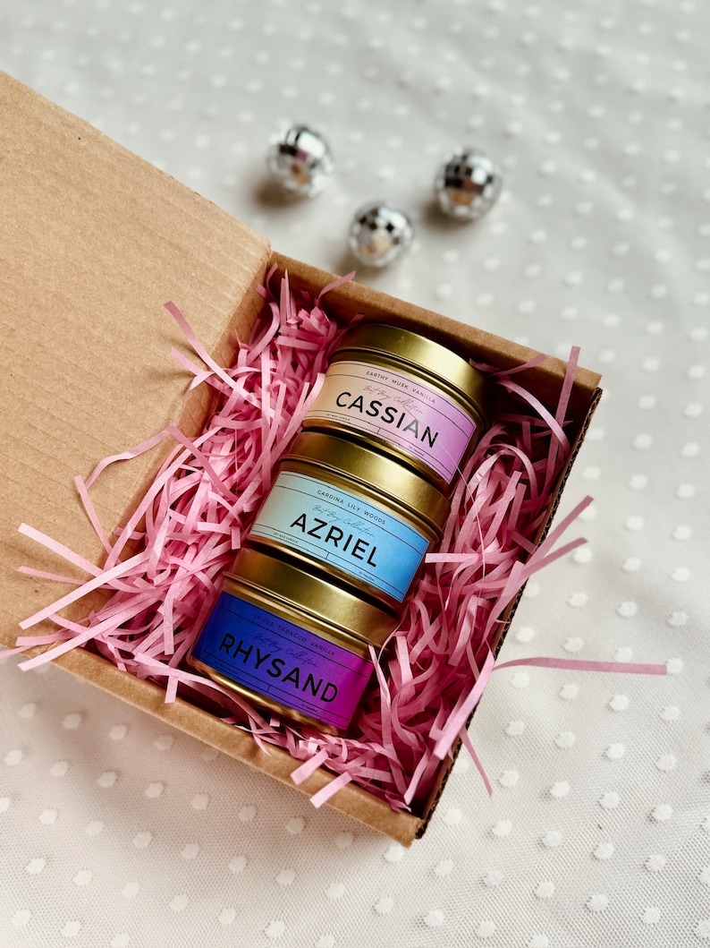 May include: Three gold candle tins with pink, blue, and purple labels. The labels have the names "Cassian", "Azriel", and "Rhysand" on them. The candles are in a brown box with pink shredded paper.