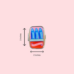 May include: A rectangular metal pill case featuring a colorful graphic of three blue fish on a pink background above a red section with the text SARDINES. The case measures 3 inches in height and 2 inches in width as indicated by the visible arrows.