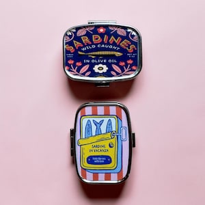 May include: Two rectangular metal tins with rounded corners. The top tin is dark blue with floral patterns and text reading SARDINES WILD CAUGHT IN OLIVE OIL. The bottom tin features red and white stripes with a yellow sardine can graphic and text SARDINE IN VACANZA from the sea with love.
