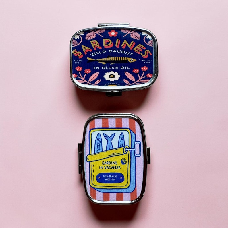 May include: Two rectangular metal tins with rounded corners. The top tin is dark blue with floral patterns and text reading SARDINES WILD CAUGHT IN OLIVE OIL. The bottom tin features red and white stripes with a yellow sardine can graphic and text SARDINE IN VACANZA from the sea with love.