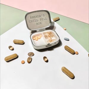 May include: A silver-toned metal pill box with a hinged lid featuring the engraved text AMANDA'S CHILL PILL BOX. The interior is divided into two compartments containing small round orange tablets and translucent oval capsules, with loose capsules scattered around.