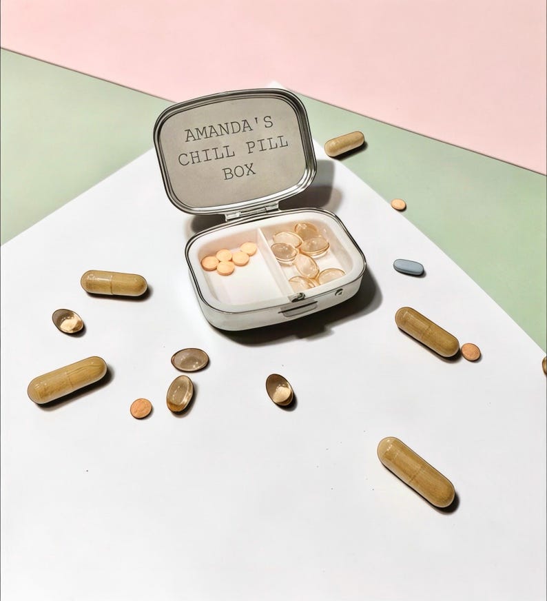 May include: A silver-toned metal pill box with a hinged lid featuring the engraved text AMANDA'S CHILL PILL BOX. The interior is divided into two compartments containing small round orange tablets and translucent oval capsules, with loose capsules scattered around.