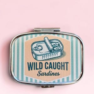May include: A rectangular metal pill box with a hinged lid featuring a graphic of an open tin of sardines. The design includes light blue and white vertical stripes, a peach-colored center panel, and the text WILD CAUGHT Sardines.