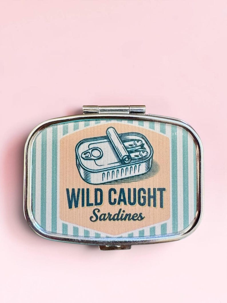 May include: A rectangular metal pill box with a hinged lid featuring a graphic of an open tin of sardines. The design includes light blue and white vertical stripes, a peach-colored center panel, and the text WILD CAUGHT Sardines.
