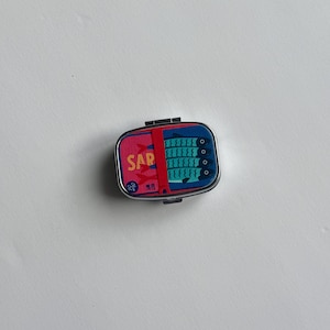 K&ouml;nnte beinhalten: Kleine, rechteckige Metallbox mit Klappdeckel. Die Box zeigt ein farbenfrohes Design mit dem Wort &bdquo;SAR&ldquo; in Rosa und einer Grafik von Sardinen in Blau. Die Box dient wahrscheinlich zur Aufbewahrung kleiner Gegenst&auml;nde.