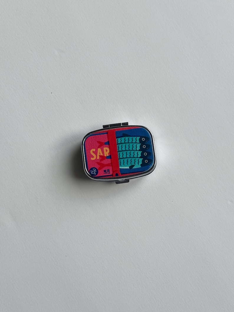 K&ouml;nnte beinhalten: Kleine, rechteckige Metallbox mit Klappdeckel. Die Box zeigt ein farbenfrohes Design mit dem Wort &bdquo;SAR&ldquo; in Rosa und einer Grafik von Sardinen in Blau. Die Box dient wahrscheinlich zur Aufbewahrung kleiner Gegenst&auml;nde.
