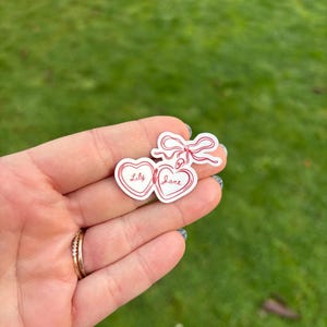 K&ouml;nnte beinhalten: Ein Aufkleber mit zwei verbundenen Herzformen, die die Namen "Lily" und "Jane" in Schreibschrift tragen. Die Herzen sind rot umrandet und haben eine rote Schleife. Der Aufkleber wird vor einem gr&uuml;nen Hintergrund gehalten.