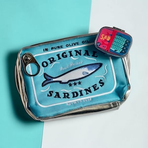 Könnte beinhalten: Eine rechteckige hellblaue Tasche im Design einer Sardinendose mit der Aufschrift "ORIGINAL SARDINES" und "IN PURE OLIVE OIL" sowie einem Metall-Ring als Akzent. Ein kleinerer, passender dosenartiger Behälter liegt oben auf.
