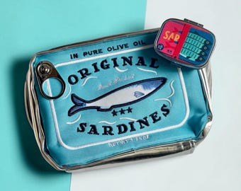 Puede incluir: Un estuche rectangular de color azul claro diseñado como una lata de sardinas, con el texto "ORIGINAL SARDINES" e "IN PURE OLIVE OIL" y un detalle de anilla metálica. Un recipiente pequeño a juego con forma de lata descansa encima.