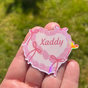 May include: Pink heart-shaped sticker with a white background and the word "Xaddy" written in pink cursive. The sticker has a holographic finish and is decorated with pink bows.