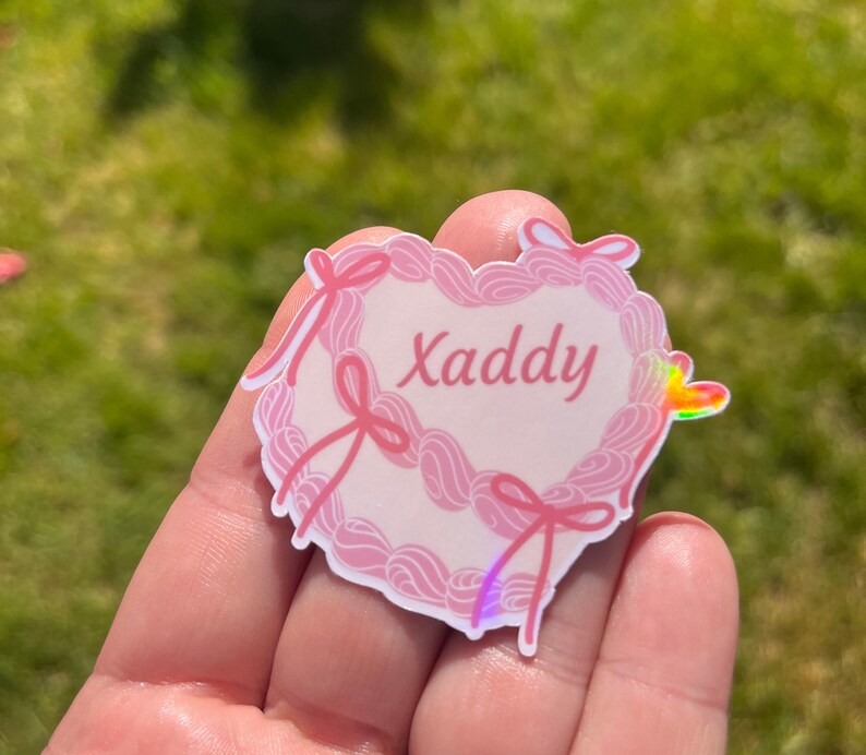 May include: Pink heart-shaped sticker with a white background and the word "Xaddy" written in pink cursive. The sticker has a holographic finish and is decorated with pink bows.