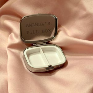 K&ouml;nnte beinhalten: Ge&ouml;ffnete, silberfarbene Pillendose mit wei&szlig;em Innenraum, der in zwei F&auml;cher unterteilt ist. Der Deckel tr&auml;gt die Gravur "AMANDA'S PILL BOX". Die Dose ist rechteckig mit abgerundeten Ecken, vor rosa Satin-Hintergrund.