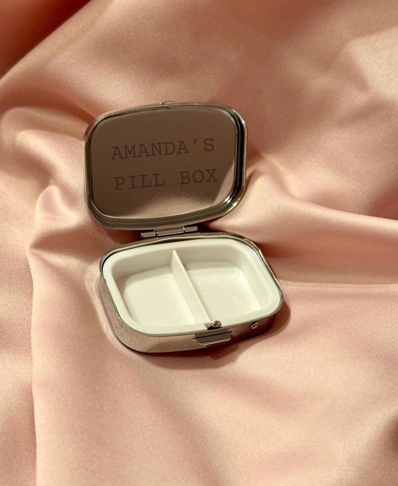 K&ouml;nnte beinhalten: Ge&ouml;ffnete, silberfarbene Pillendose mit wei&szlig;em Innenraum, der in zwei F&auml;cher unterteilt ist. Der Deckel tr&auml;gt die Gravur "AMANDA'S PILL BOX". Die Dose ist rechteckig mit abgerundeten Ecken, vor rosa Satin-Hintergrund.