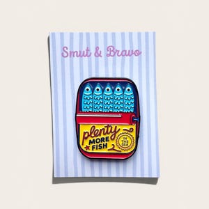May include: A rectangular enamel pin featuring a sardine tin design with five blue fish inside. The yellow label reads "plenty MORE FISH" with a small red star and a circular badge stating "IN THE SEA". The pin is attached to a light blue striped card.