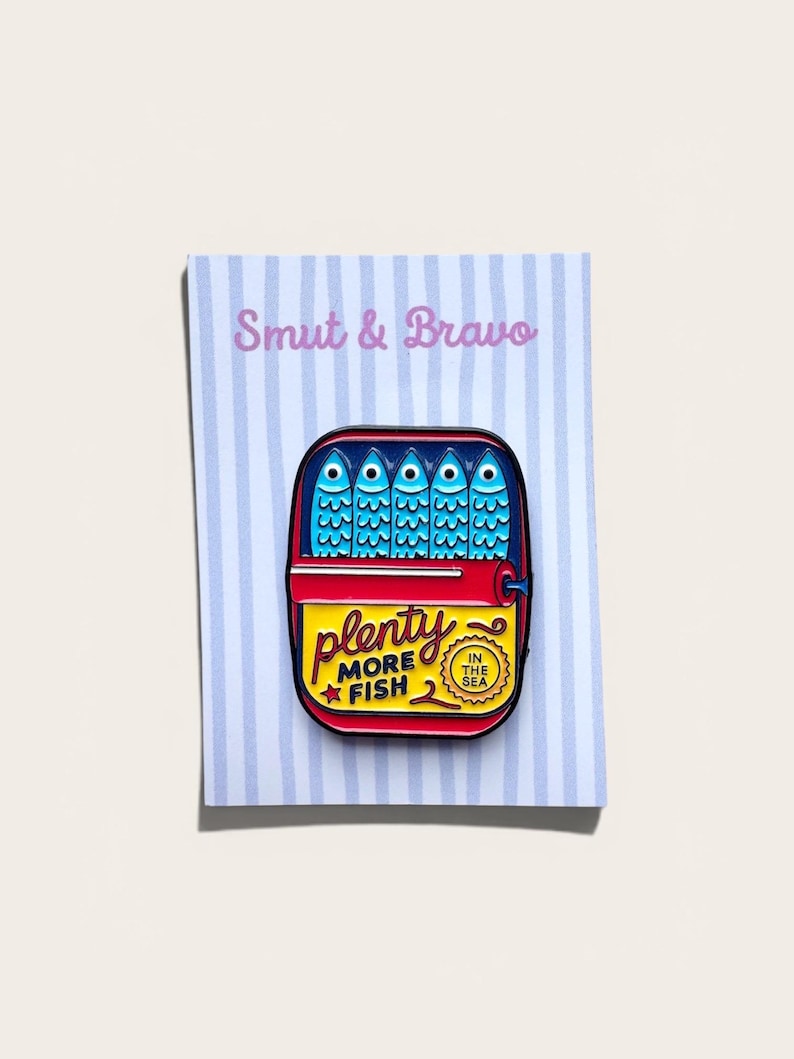 May include: A rectangular enamel pin featuring a sardine tin design with five blue fish inside. The yellow label reads "plenty MORE FISH" with a small red star and a circular badge stating "IN THE SEA". The pin is attached to a light blue striped card.